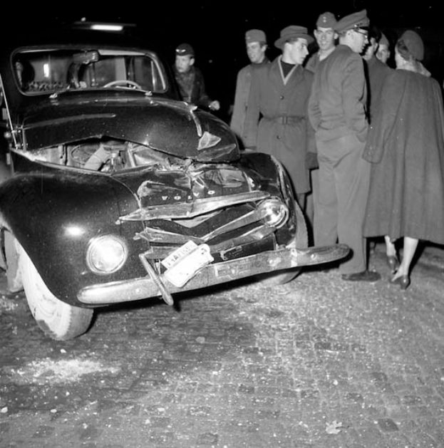 Ein Schwarz-Weiß-Bild eines beschädigten Autos am Straßenrand, umgeben von einer Gruppe besorgter Menschen