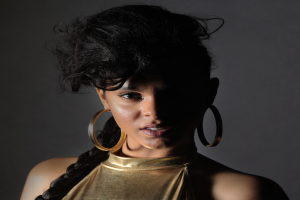 Eine Frau in einem goldenen Kleid und großen Kreolen steht selbstbewusst vor einer Wand, ihr glatter Bob und natürliches Make-up unterstreichen ihren entschlossenen Ausdruck, während sie direkt in die Kamera blickt.