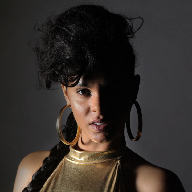 Eine Frau in einem goldenen Kleid und großen Kreolen steht selbstbewusst vor einer Wand, ihr glatter Bob und natürliches Make-up unterstreichen ihren entschlossenen Ausdruck, während sie direkt in die Kamera blickt.