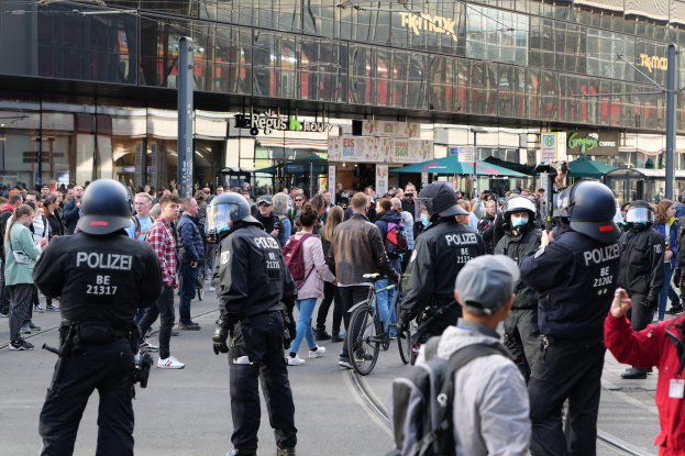 Eine Gruppe von Polizeibeamten vor einer Menschenmenge auf der Straße stehend, wobei einige Personen Helme tragen und Taschen oder Fahrräder tragen, und Gebäude mit Glaswänden und Schildern im Hintergrund.