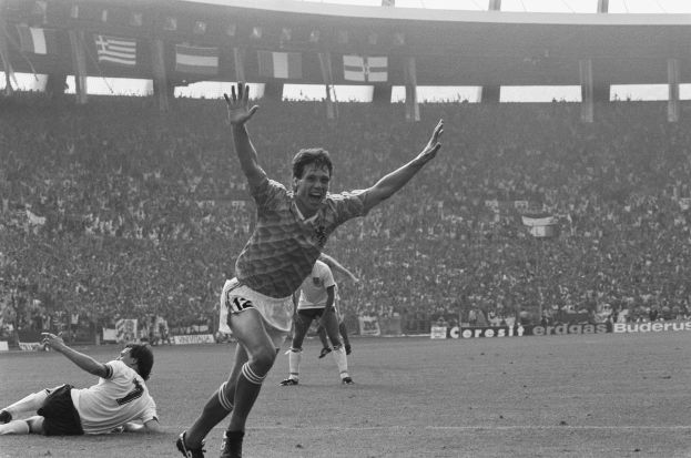 Ein Schwarz-Weiß-Foto eines Fußballspielers, der nach einem Tor feiert, umgeben von Menschen auf dem Boden, mit Schildern, Fahnen und Pfosten im Hintergrund unter einem klaren Himmel.