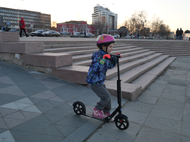 Ein junges Mädchen in einem Helm fährt mit einem Scooter eine Treppe hinunter, mit Fahrzeugen, Menschen, Laternen, Bäumen, Gebäuden und einem klaren blauen Himmel im Hintergrund.