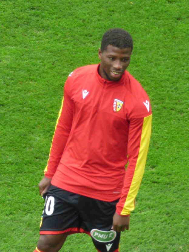 Ein Mann in einem roten T-Shirt und schwarzen Shorts, der auf einem Fußballfeld steht und lächelt.