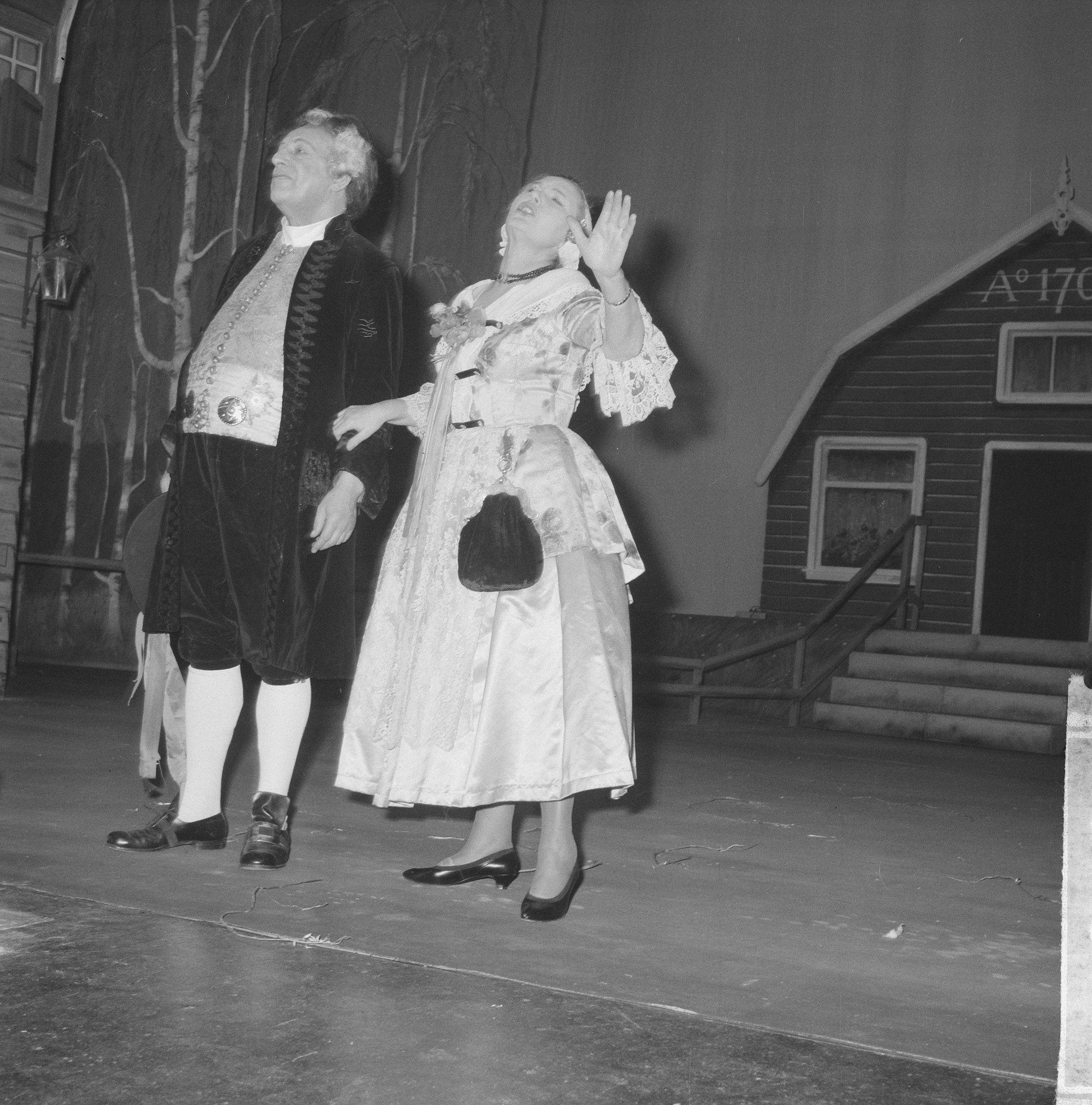 Ein Mann in einem schwarz-weißen Kleid und eine Frau stehen auf einer Bühne vor einem Haus, mit Treppe, Geländer, Wand, Laterne und Bäumen im Hintergrund.