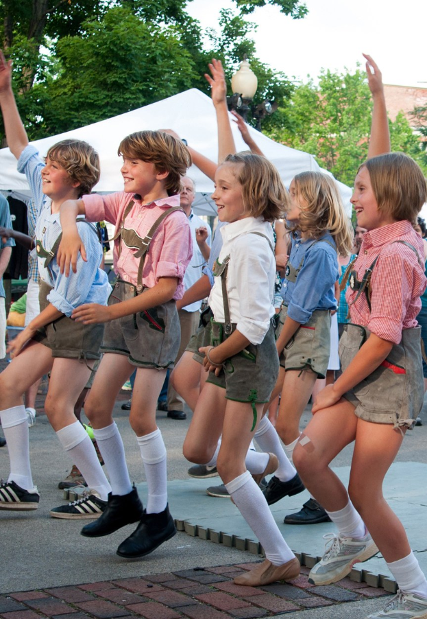 Kinder in traditioneller bayrischer Tracht tanzen auf einer Straße bei einem Outdoor-Festival, umgeben von einer Menge, Zelten und Gebäuden unter einem klaren blauen Himmel.