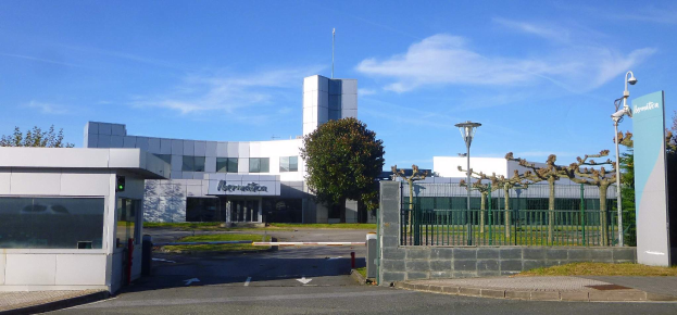 Ein großes, mehrfenstriges Gebäude mit einem Tor davor, umgeben von Straßenmasten, Laternen, Gittern, Bäumen, Gras und einem bewölkten Himmel, das als Hauptsitz von Iberostica in den Niederlanden dient.