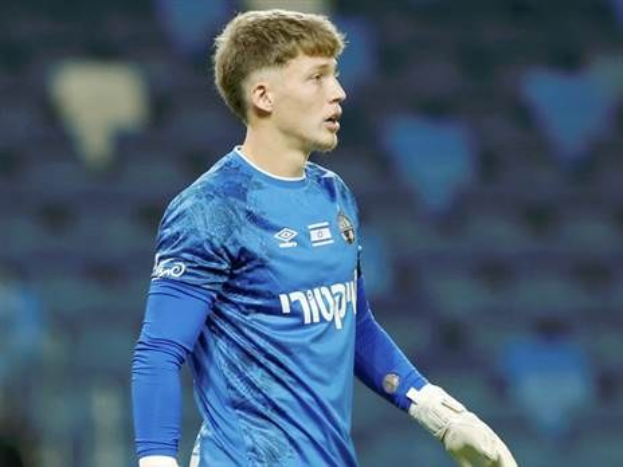 Ein junger Mann in einem blauen Torwarttrikot steht auf einem Fussballfeld und trägt ein blaues T-Shirt und weisse Handschuhe während eines Spiels.