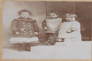 Drei Kinder auf altem Schwarz-Weiß-Foto stehen eng zusammen, Gesichter zeigen Trauer, mit einer Wand dahinter.