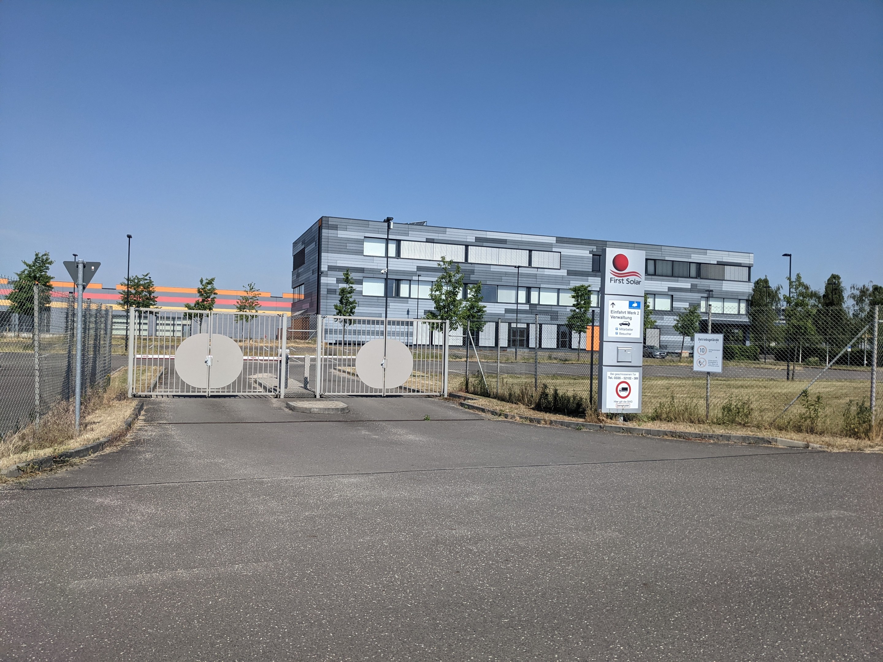 Großes industrielles Gebäude mit der Aufschrift "First Solar" und zahlreichen Fenstern, umgeben von einem Metallzaun, Bäumen, Pflanzen, Gras, Schildern, Straßenmasten und einem Weg.