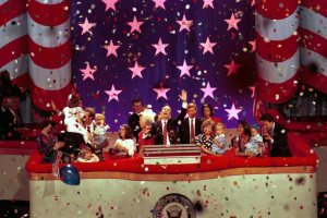 Eine Gruppe von Menschen steht auf einer Bühne mit einem Rednerpult, das ein Logo, Luftballons und Konfetti im Hintergrund zeigt.