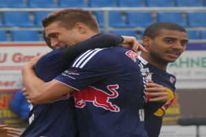 Zwei Fußballspieler umarmen sich auf einem Feld, mit einer Hand auf der linken Seite sichtbar und im Hintergrund einige Menschen, Schilder mit Text und blaue Stühle.