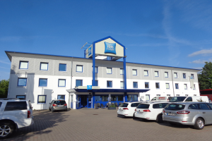 Gebäude mit "ibis budget frankfurt airport"-Schilder, geparkten Autos, Topfpflanzen, einer Straßenlaterne, Bäumen und einem bewölkten Himmel.