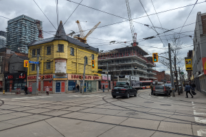 Eine Straßenszene in der Stadt mit einem gelben Gebäude im Bau, Autos, Fußgängern, Gehwegen, Strommasten mit Drähten, Ampeln, Gebäuden mit Fenstern, Schildern, Bäumen und einem bewölkten Himmel.