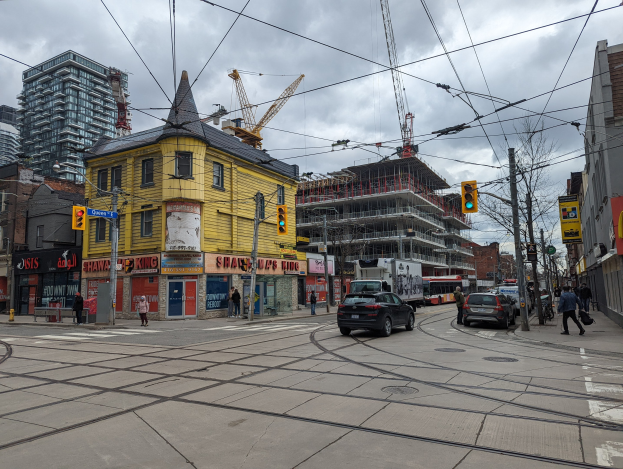 Eine Straßenszene in der Stadt mit einem gelben Gebäude im Bau, Autos, Fußgängern, Gehwegen, Strommasten mit Drähten, Ampeln, Gebäuden mit Fenstern, Schildern, Bäumen und einem bewölkten Himmel.