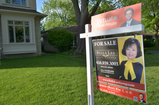 Immobilie zum Verkauf Schild vor einem Haus mit Fenstern, umgeben von grünem Gras, Bäumen und Pflanzen unter einem sichtbaren Himmel.