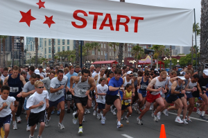Gruppe von Läufern bei einem Marathon mit einem Verkehrskegel im Vordergrund und einer Fahne im Hintergrund, umgeben von Bäumen, Laternenmasten, Gebäuden und einem klaren blauen Himmel.