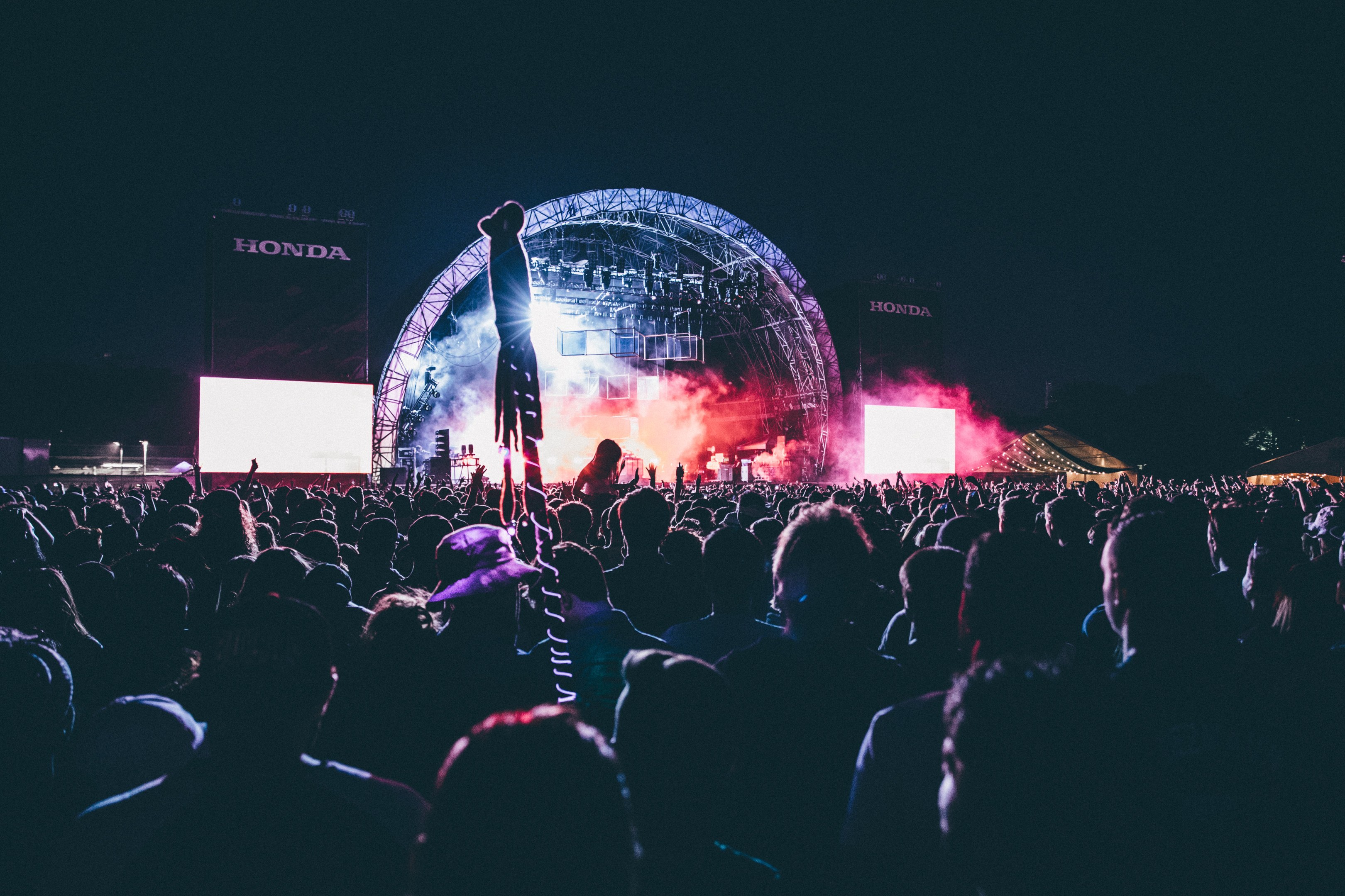 Eine große Menge Menschen auf einem Musikfestival mit einem Künstler auf einer hell erleuchteten Bühne umgeben von Musikern, Bildschirmen und Bannern in einer dunklen Umgebung.