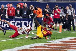 Ein Football-Spieler taucht in die Endzone für einen Touchdown ein, mit einer Gruppe von Menschen am Boden, einige halten Kameras, und eine Fahne mit Text im Vordergrund und Geländern und Pfosten im Hintergrund.