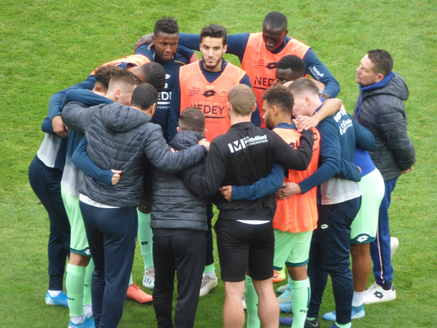 Eine Gruppe von M%C3%A4nnern in Jacken stand auf einem Fussballfeld im Kreis.