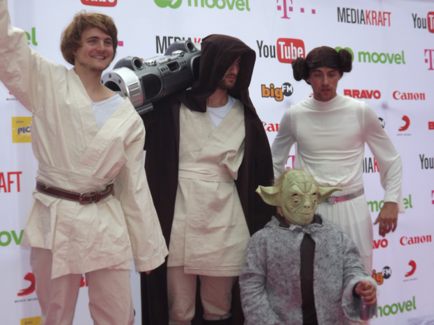 Gruppe von Menschen in Star-Wars-Kostümen, die auf einem roten Teppich posieren und im Hintergrund ein Banner mit Text und Logos zu sehen ist.