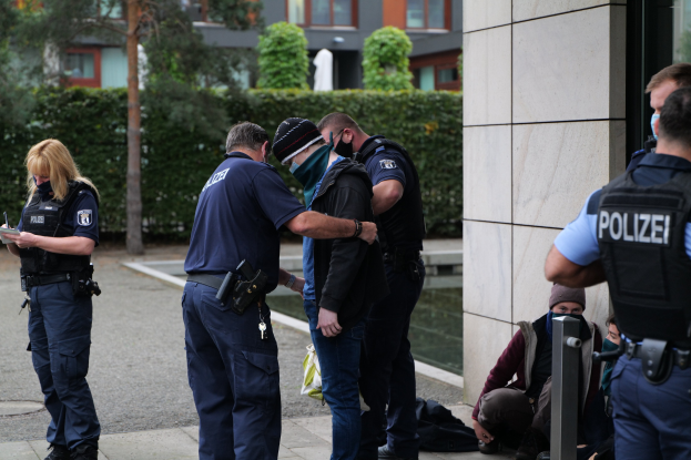 Gruppe von Polizeibeamten vor einem Gebäude mit einer Frau, die ein Buch und einen Stift hält, während andere sitzen oder stehen, mit Bäumen und Gebäuden im Hintergrund.