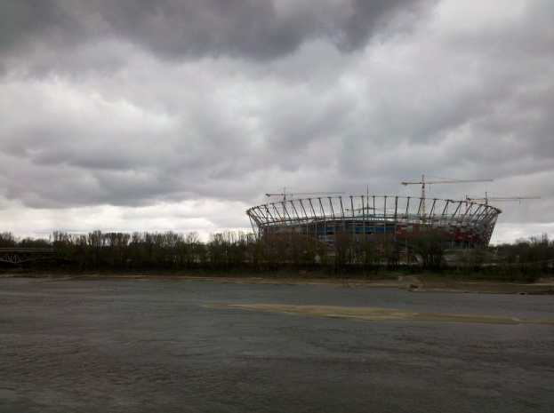 Das Olympiastadion in Kiew, Ukraine, ein großes Stadion umgeben von Bäumen und einer Brücke, mit einem bewölkten Himmel im Hintergrund und dem Boden unten.