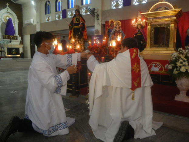 Zwei Priester knien vor einer Jesus-Statue, umgeben von brennenden Kerzen, mit Blumenschmuck auf beiden Seiten des Altars und Menschen im Hintergrund.