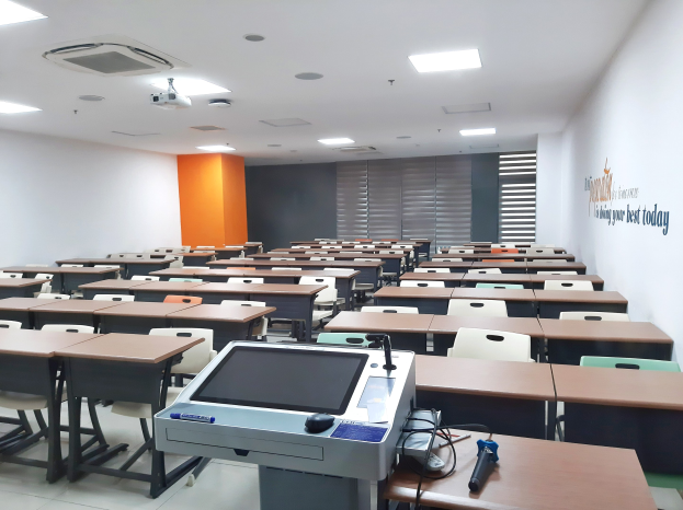 Ein Klassenzimmer mit Tischen und Stühlen in Reihen, ein deckenmontierter Projektor und Gegenstände auf Tischen, mit Text an der rechten Wand, der auf eine Privatschule in Singapur hinweist.