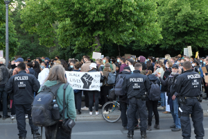 Eine große Gruppe von Menschen steht an der Straße, einige halten Schilder, mit einem Fahrrad im Vordergrund und Bäumen und einem Pfahl im Hintergrund, bei einer Black-Lives-Matter-Demonstration in Berlin.
