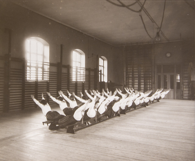 Menschen, die Pilates-Übungen in einem Fitnessstudio mit einer Wanduhr und Fenstern im Hintergrund durchführen.