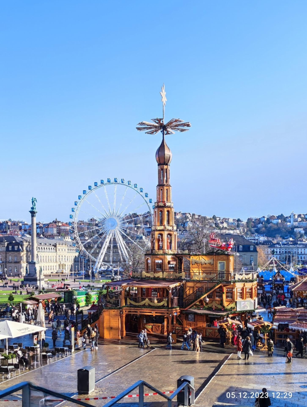 Ein belebter Stadtplatz mit einem zentralen Riesenrad, umgeben von Gebäuden, Bäumen und Menschen unter einem klaren blauen Himmel, mit Zelten und Geländern im Bild und einem Wasserzeichen, das es als einen der besten Weihnachtsmärkte in Europa ausweist.