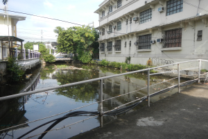 Kleiner Kanal mit Geländern, Rohren und Grünpflanzen, der zwischen hohen Gebäuden in einer städtischen Umgebung verläuft, mit Stromleitungen und einem bewölkten Himmel.