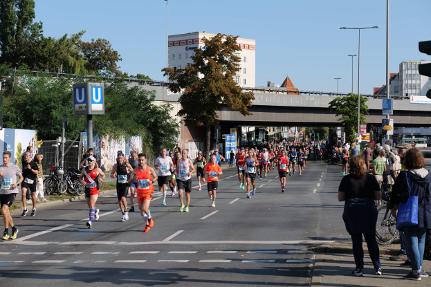 Gruppe von Menschen, die einen Marathon auf einer von Bäumen gesäumten Straße mit Fahrrädern, Schildern, einem Zaun, einer Brücke, Gebäuden und einem klaren blauen Himmel laufen.