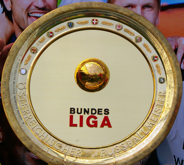Ein Mann hält einen goldenen Schild mit 'Bundesliga' darauf, im Hintergrund eine Fahne mit drei Personen.