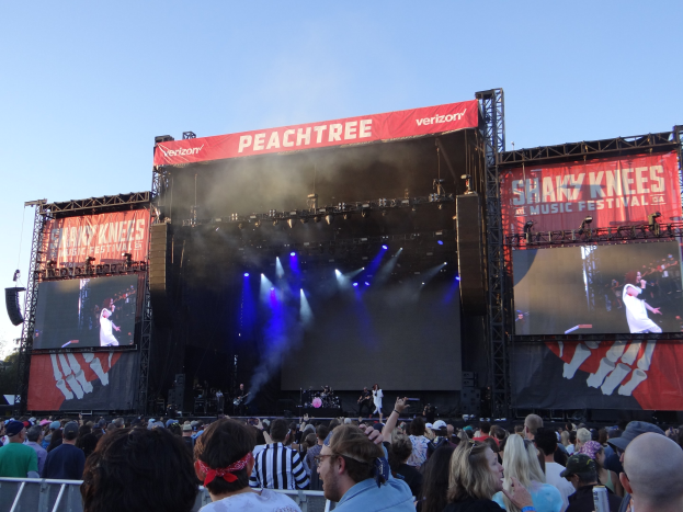 Eine große Menschenmenge steht vor einer hell erleuchteten Bühne auf dem Shaky Knees Music Festival, wo Musiker unter Bannern mit Text spielen, mit einem Geländer im Vordergrund und einem klaren blauen Himmel im Hintergrund.