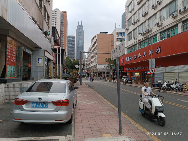 Ein Mann fährt mit einem Roller eine Straße in der Stadt entlang, gesäumt von hohen Gebäuden, mit Menschen auf den Bürgersteigen, Fahrzeugen auf der Straße und Bäumen unter einem klaren blauen Himmel im Hintergrund.