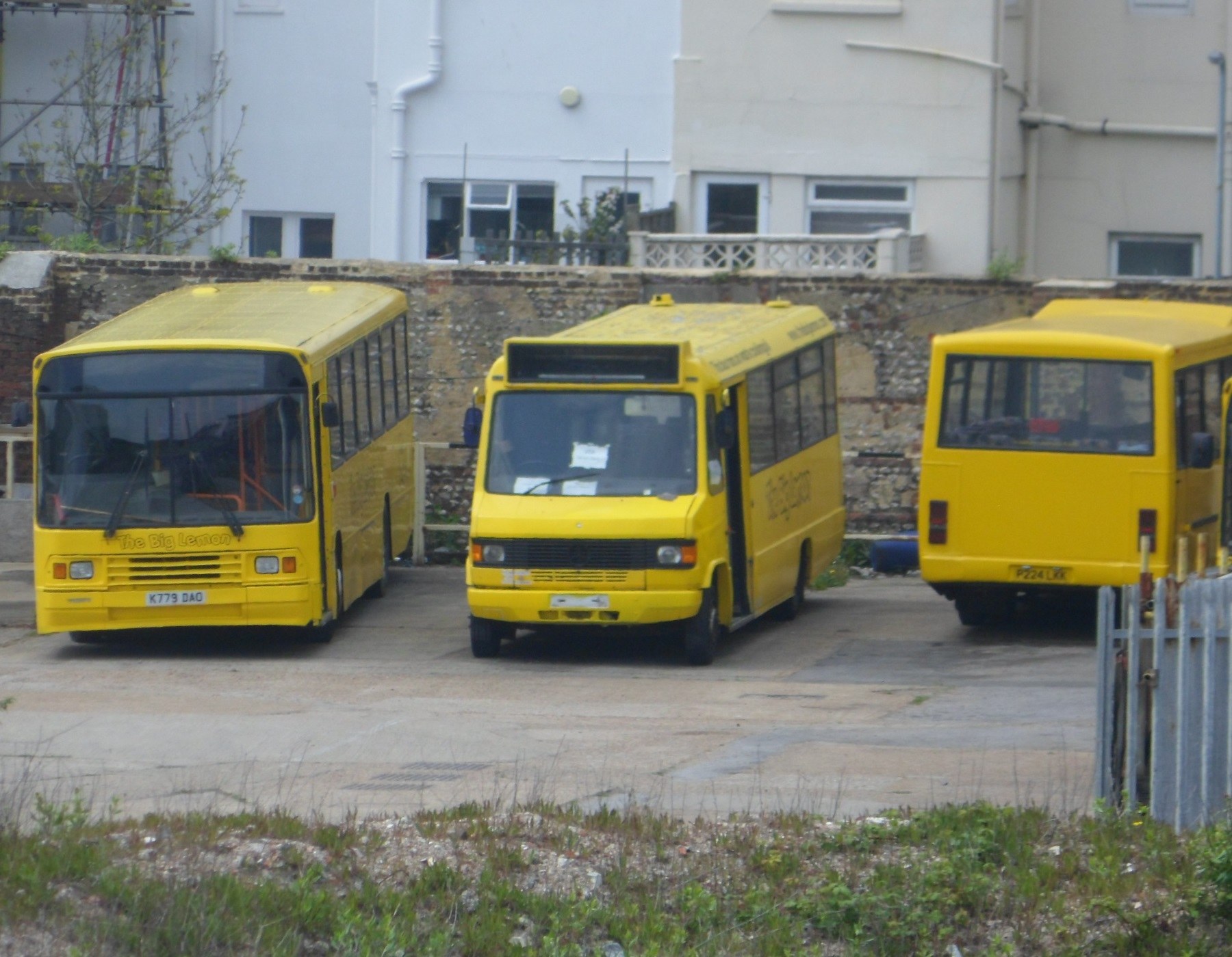 Drei gelbe Busse nebeneinander auf einem eingezäunten Parkplatz mit Grünfläche, einem Gebäude und Versorgungsanlagen.