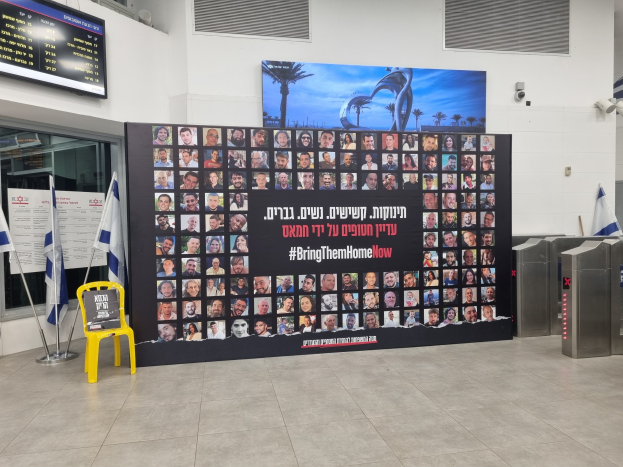 Eine große Wand zeigt zahlreiche Fotos, einen gelben Stuhl, israelische Regierungsflaggen auf Ständern, eine Tafel mit dem Text "Bring Them Home Now", einen montierten Bildschirm, Deckenleuchten und andere Gegenstände.