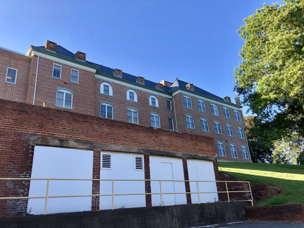 Großes Backsteingebäude mit zahlreichen Fenstern, umgeben von einem Metallzaun, Weg, Gras, Bäumen und einem bewölkten Himmel, auf dem Campus der University of Virginia gelegen.