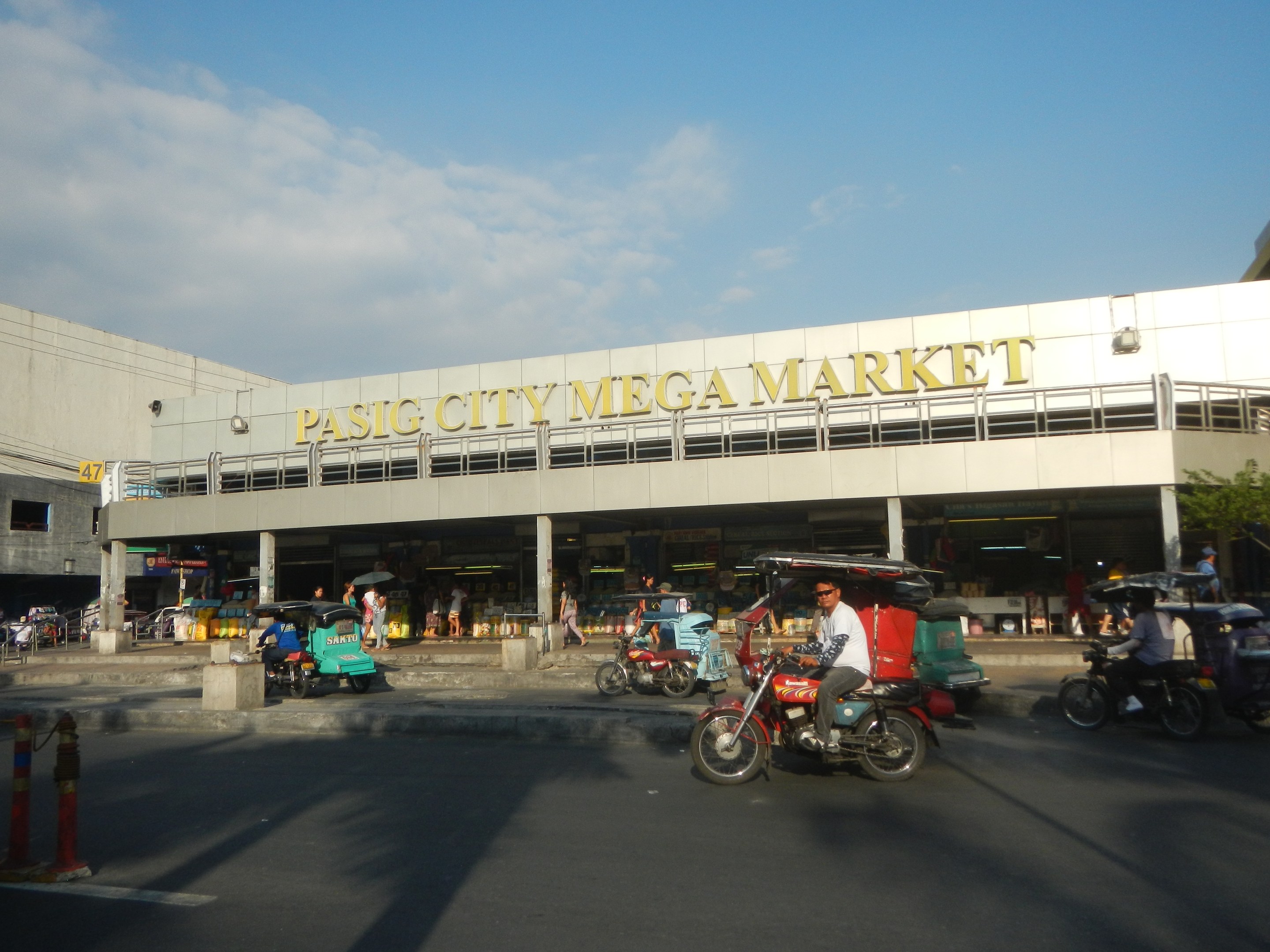 Ein belebter Pasig City Mega Market mit Fahrzeugen auf der Straße, Menschen, die herumlaufen, einem Gebäude mit Text im Hintergrund und einem bewölkten Himmel.