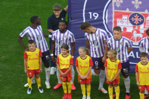 Eine Gruppe junger Menschen auf einem saftig grünen Fußballfeld, im Hintergrund ein Banner mit der Aufschrift "Ligue 1", einige tragen Trikots und andere Shorts, unter einem strahlend blauen Himmel.