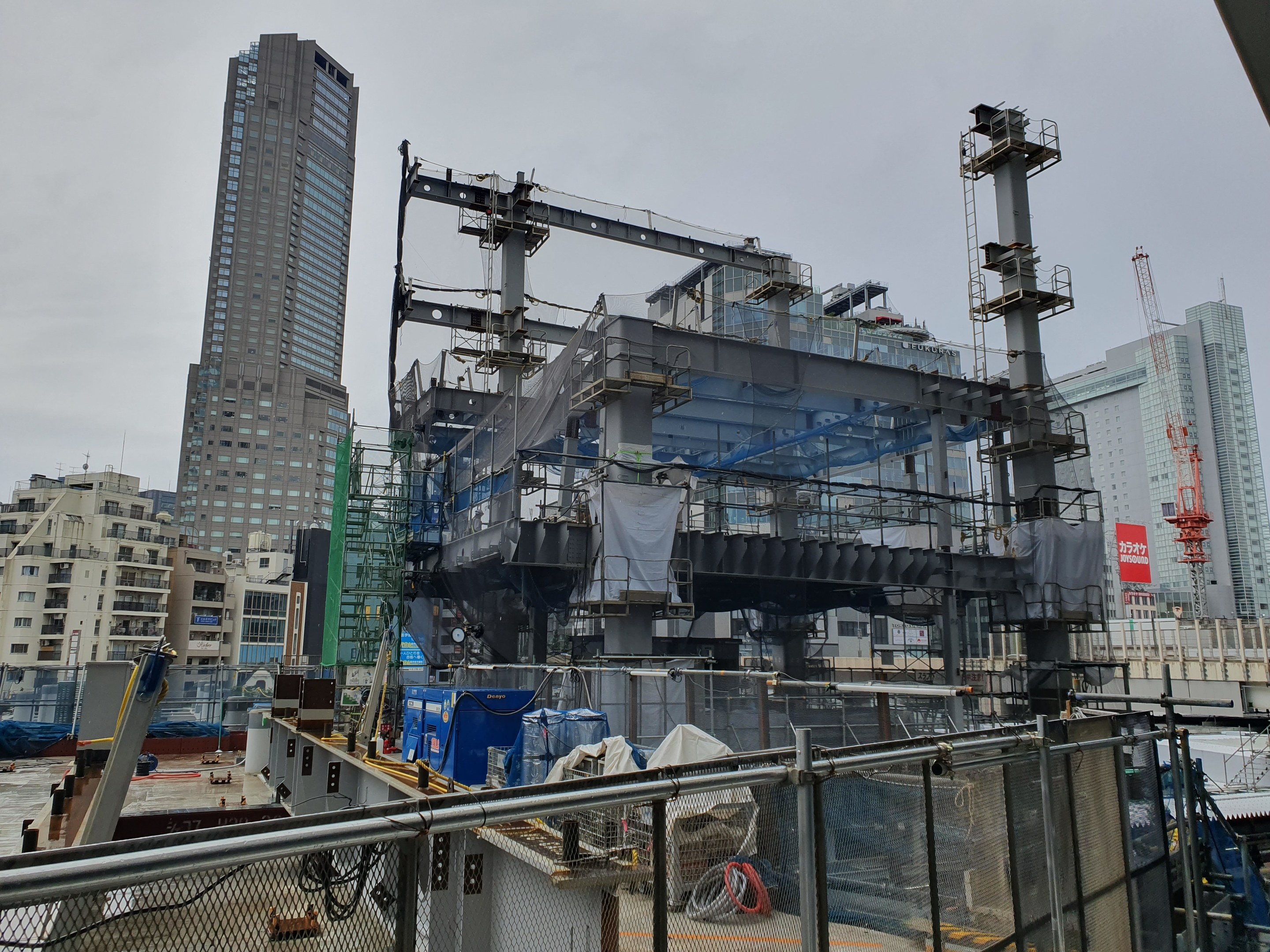 Großbaustelle in einer Stadt mit einem teilweise errichteten Kraftwerk, umgeben von einem Metallzaun, umgeben von Gebäuden, Fahrzeugen, Pfählen, Drähten und einem bewölkten Himmel.