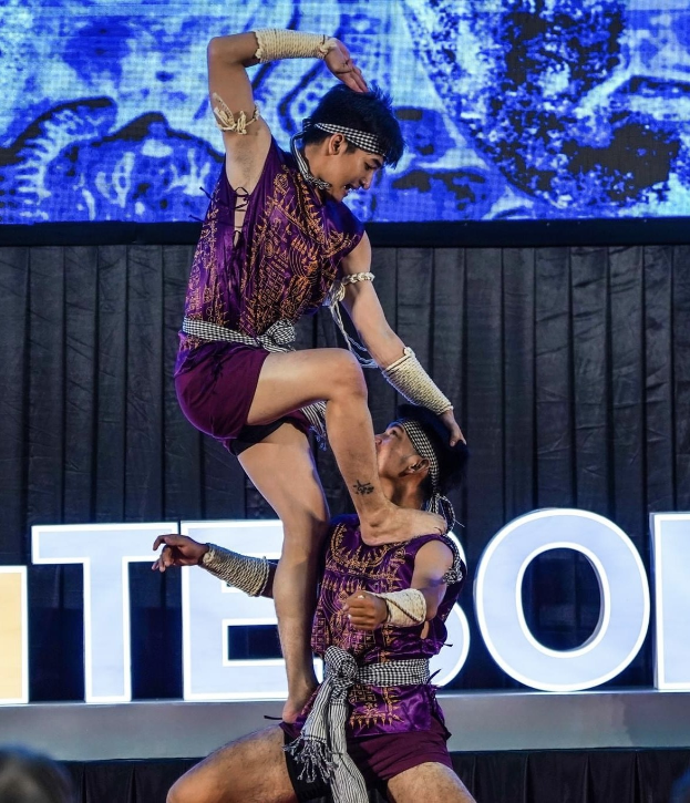 Zwei Performer in purpurfarbenen Kleidern führen einen akrobatischen Hebemanöver auf der Bühne vor einem Publikum aus, mit einem schwarzen Vorhang und einer Bildschirmrückwand.