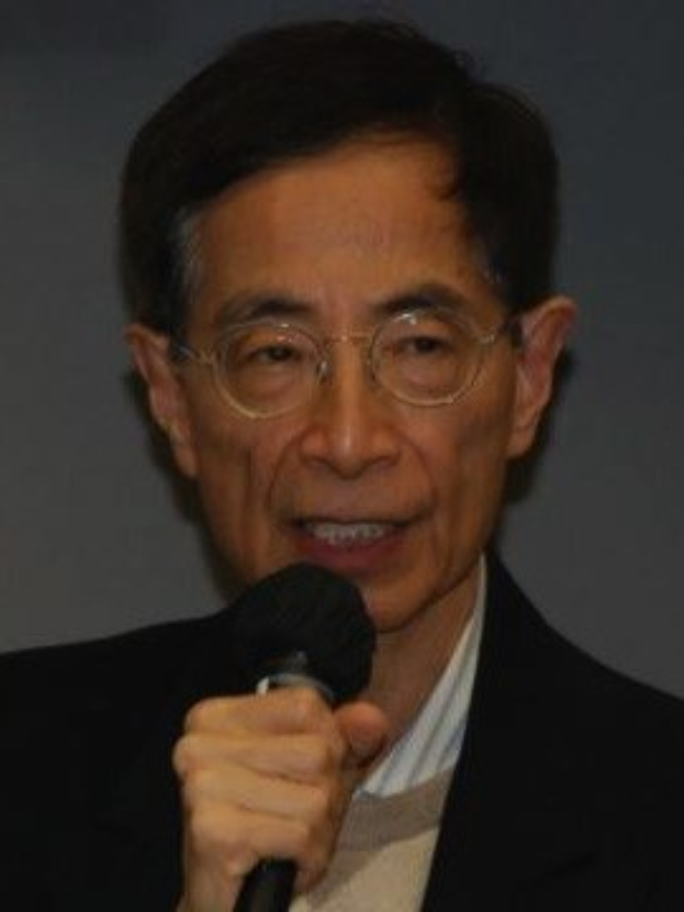 Ein Mann in einem schwarzen Blazer und Brillengläsern hält ein Mikrofon und sieht ernst aus, wahrscheinlich als Ökonom.