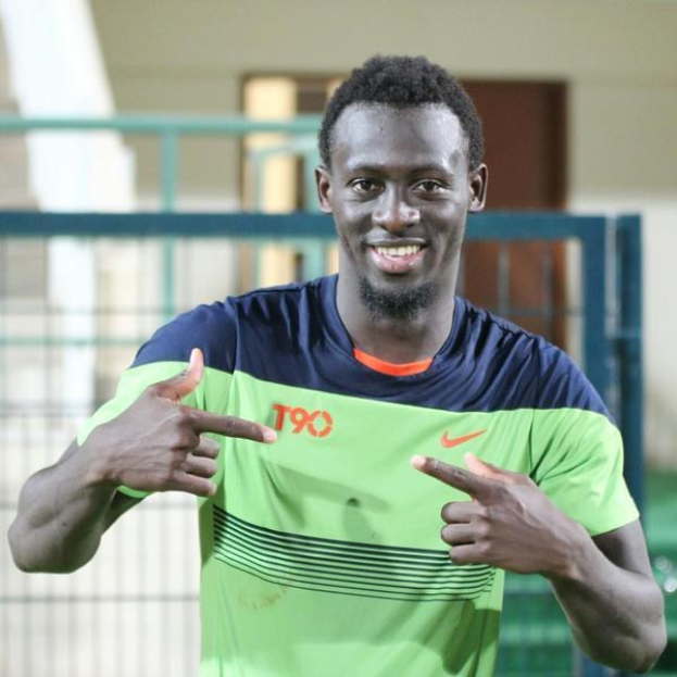 Ein Mann in einem grün-blauen T-Shirt, wahrscheinlich Emmanuel Adebayor, zeigt mit einem Lächeln auf die Kamera, mit einem unscharfen Geländer, Wand und Tür im Hintergrund.