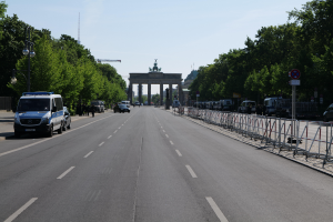 Ein Polizeifahrzeug steht auf der Seite einer vielbefahrenen Straße vor dem Brandenburger Tor in Berlin, Deutschland, mit Barrieren, Schildern, Bäumen und Laternen im Hintergrund und einem bewölkten Himmel.