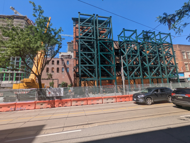 Große Metallbrücke im Bau an einer Baustelle mit parkenden Autos, einem Zaun, Bäumen, Gebäuden, einem Kran und dem Himmel im Hintergrund.
