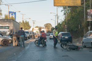 Eine Gruppe von Menschen steht um ein verunglücktes Motorrad auf der Straße mit mehreren Fahrzeugen, darunter ein Lastwagen, und einem Hintergrund aus Bäumen, Polen, Lampen und Schildern unter dem Himmel.