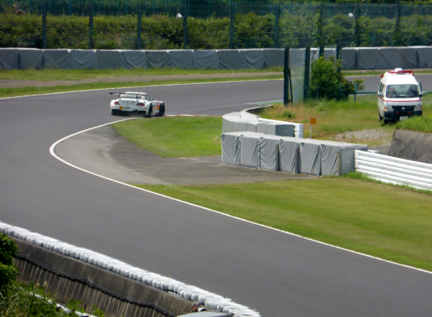 Ein Rennwagen fährt eine Rennstrecke entlang, im Hintergrund ist ein Polizeiauto zu sehen, umgeben von Gras, Pflanzen, Bäumen und einem Zaun, mit einem Fahrzeug auf der rechten Seite und Text unten.