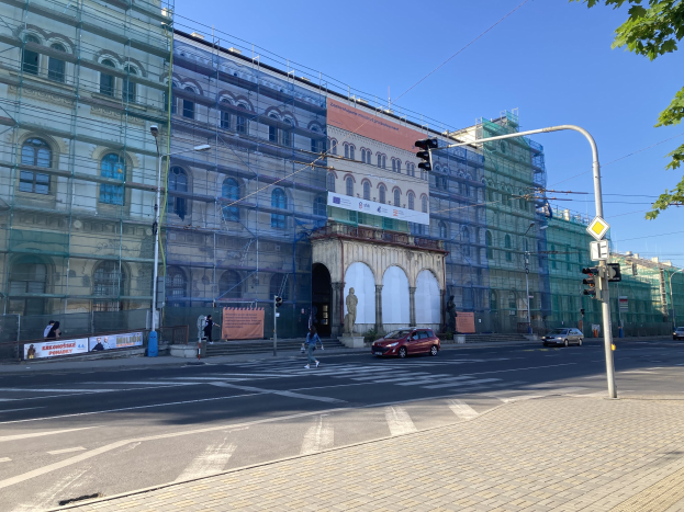 Ein großes Gebäude mit Gerüst, umgeben von Fahrzeugen, Fußgängern und städtischer Infrastruktur, unter einem klaren blauen Himmel.